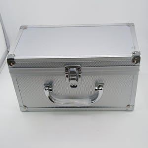 May include: A rectangular silver storage case with a textured diamond pattern finish. The case features reinforced metal corners, a central silver latch mechanism, and a sturdy curved handle with a protective plastic wrapping on the grip.