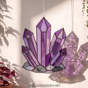 May include: A stained glass amethyst crystal cluster in shades of purple, lavender, and white. The suncatcher is suspended by a thin wire, casting a shadow on the wall. The base is a grey, textured stone. The website address is visible at the bottom.