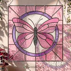 May include: A stained glass panel with a butterfly design in pink and purple hues. The butterfly is centrally positioned within a circular frame, enclosed by a square border. The artwork is suspended by a chain.