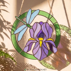May include: A circular stained glass suncatcher featuring a purple iris flower with a yellow center and a light blue dragonfly. The design is framed by a green border and includes a jute twine hanger for display, with the text glassgarden.net visible.