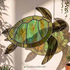 May include: A stained glass sea turtle suncatcher featuring a shell with marbled green and amber glass panels held together by dark metal solder lines. The turtle is suspended by a silver chain and displays the text GLASSGARDEN.NET at the bottom.