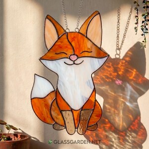 Può includere: Suncatcher in vetro colorato a forma di volpe sorridente, nei toni dell'arancione, bianco e marrone. La volpe è appesa a una catena d'argento. Il testo "GLASGARDEN.NET" è visibile in basso.