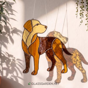 Op de afbeelding: Gebrandschilderde glazen zonnevanger van een hond in bruin- en goudtinten. De hond is afgebeeld in een staande pose, met een gedetailleerd ontwerp en zichtbare loodlijnen. De zonnevanger hangt aan een dunne draad. De website GLASSGARDEN.NET is zichtbaar.
