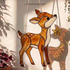 May include: Stained glass deer with a star. The deer is brown, tan, and white with a star hanging from its mouth. The deer has white spots on its back. The image includes the text "glassgarden.net".