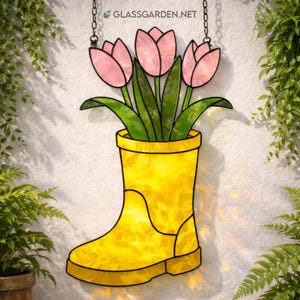 May include: A stained glass artwork depicting a yellow wellington boot filled with pink tulips and green leaves. The piece is suspended by a chain and set against a white wall with foliage. The website address "GLASSGARDEN.NET" is visible.