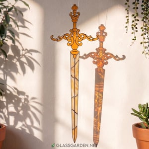 May include: A decorative stained glass sword with an ornate gold-colored hilt and a light-colored blade. The sword is suspended in front of a white wall, casting a shadow. The text "GLASSGARDEN.NET" is visible at the bottom.