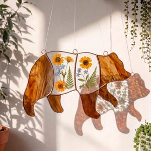 May include: A stained glass bear suncatcher with a copper-coloured frame. The bear's body is divided into sections, with pressed yellow sunflowers, blue forget-me-nots, and green ferns. The bear is hanging from a white string.