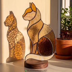 Könnte beinhalten: Eine Buntglas-Skulptur in Katzenform, bestehend aus verschiedenen bernsteinfarbenen, braunen und cremefarbenen Glasstücken, die durch dunkle Metalllötstellen verbunden sind. Die Katze steht auf einem runden Holzsockel und wirft einen Schatten.
