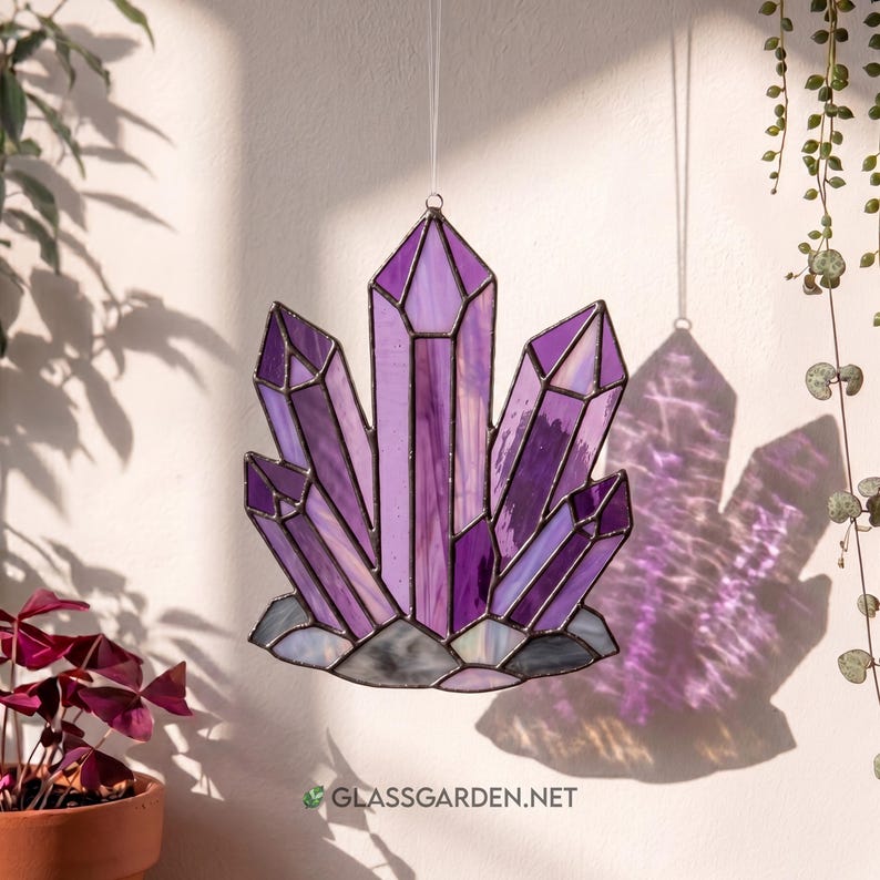 K&ouml;nnte beinhalten: Ein Buntglas-Amethyst-Kristallcluster in Lila-, Lavendel- und Wei&szlig;t&ouml;nen. Der Sonnenf&auml;nger wird von einem d&uuml;nnen Draht gehalten und wirft einen Schatten an die Wand. Die Basis ist ein grauer, strukturierter Stein. Die Website-Adresse ist unten sichtbar.
