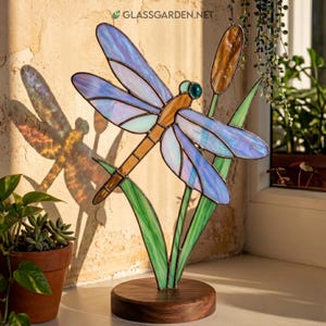 Puede incluir: Una escultura de libélula de vidriera con alas azules y moradas iridiscentes, cuerpo marrón y ojos verdes. La libélula descansa sobre hojas de vidrio verde y una espadaña marrón, montada sobre una base de madera redonda. El sitio web GLASSGARDEN.NET es visible.