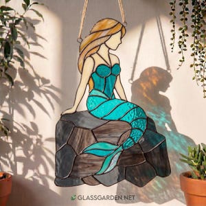 Op de afbeelding: Gebrandschilderde glazen zeemeerminfiguur in tinten van teal, goud en bruin, zittend op een rots. De zeemeermin heeft blond haar en wordt opgehangen door een jute touw. De website glassgarden.net is zichtbaar.