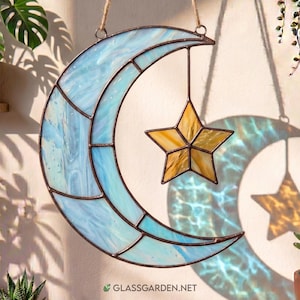 Op de afbeelding: Gebrandschilderde maan- en ster-suncatcher. De maan is lichtblauw met witte wervelingen en de ster is geel en goud. De suncatcher hangt aan een natuurlijke vezelkoord. De website glassgarden.net is zichtbaar.