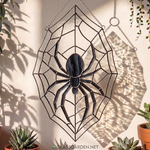 Peut inclure: Un attrape-soleil en vitrail représentant une araignée au centre d'une toile. L'objet est composé de panneaux de verre texturé transparent et de soudures en métal sombre, suspendu par une chaîne en métal argenté pour l'accrochage.