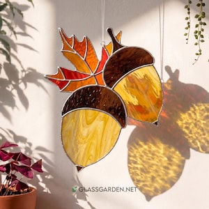 May include: Stained glass artwork featuring two acorns and a maple leaf in shades of brown, orange, and yellow. The artwork is suspended, casting a shadow on the wall. The website address "GLASSGARDEN.NET" is visible at the bottom.