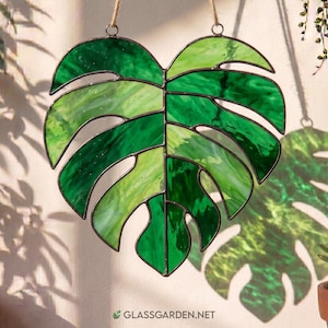 May include: A stained glass monstera leaf in shades of green, with a dark metal frame and a hanging loop. The leaf is suspended by a natural fiber cord. The website address "GLASSGARDEN.NET" is visible at the bottom.