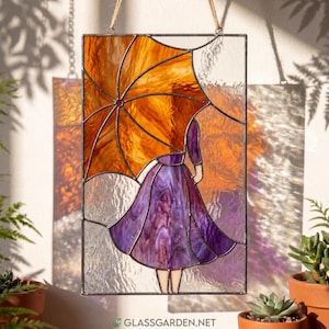 May include: Stained glass panel showing a figure with an orange umbrella and a purple dress. The artwork is rectangular, with a textured glass background. It is suspended by a natural fibre cord. The website address "glassgarden.net" is visible at the bottom.