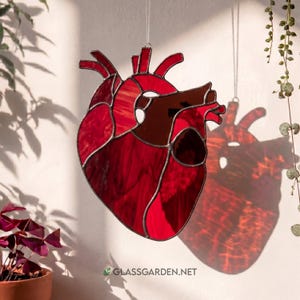 May include: A stained glass heart in shades of red and brown, with a silver-colored border. The heart-shaped artwork is suspended by a thin string. The image includes the text "GLASSGARDEN.NET".