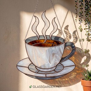 May include: Stained glass coffee cup and saucer with steam rising from the cup. The cup and saucer are white with gray and brown accents. The piece is hanging from a chain. The website address is visible at the bottom.