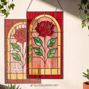 May include: A rectangular stained glass panel featuring a central red rose with green leaves, encased in an arched yellow border against a pinkish background, framed by deep red glass sections, and suspended by a metal chain for display.