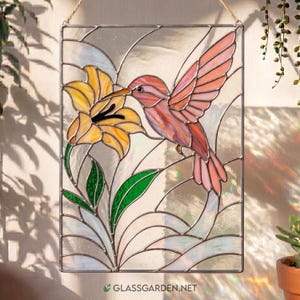 May include: Stained glass panel featuring a pink hummingbird hovering near a yellow lily. The artwork includes green leaves and is set against a clear, textured background. The panel is rectangular and hangs from a natural fiber cord. The website address is visible at the bottom.