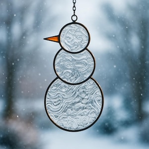 May include: A stained glass snowman ornament with a black metal frame and chain for hanging. The snowman has three circular sections, a bright orange beak, and a textured, swirling pattern within the glass. The background is a snowy, blurred scene.