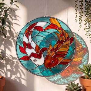 Könnte beinhalten: Ein rundes Buntglasfenster mit zwei Koi-Karpfen in einem Yin-Yang-Design. Die Fische sind rot, orange und weiß, vor einem türkisfarbenen und blauen Hintergrund. Das Fenster ist in dunklem Metall gerahmt und hängt vor einer Wand.