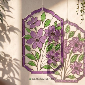 May include: A decorative stained glass panel with a floral design. The panel features purple flowers and green leaves, set within a geometric frame. The website address "glassgarden.net" is visible at the bottom. The panel is suspended, allowing light to pass through.
