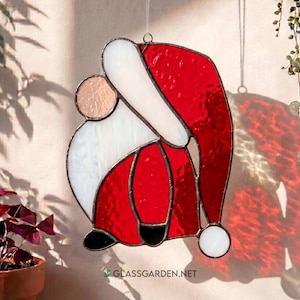 Könnte beinhalten: Glasfenster-Weihnachtsschmuck mit Santa Claus. Der Schmuck hat einen roten Hut und Anzug mit weißer Verzierung und einem runden Gesicht. Das Design ist schwarz umrandet. Der Schmuck hängt an einem silbernen Faden. Die Website-Adresse ist unten sichtbar.