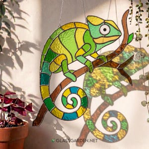 May include: A stained glass chameleon suncatcher in shades of green, yellow, and blue, perched on a brown branch. The chameleon has a curled tail and is hanging from a wire. The website address GLASSGARDEN.NET is visible at the bottom.