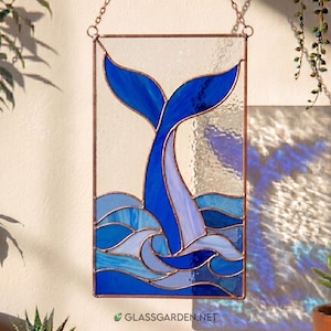 Può includere: Un acchiappasole rettangolare in vetro colorato raffigurante una coda di balena blu che emerge da onde stilizzate. Il pezzo è incorniciato in metallo color rame e appeso a una catena. Include vetro trasparente, testurizzato e blu.