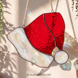 May include: A stained glass suncatcher shaped like a Santa hat, featuring vibrant red glass for the main body and iridescent white glass for the brim and pom-pom. The pieces are joined by dark metal solder and hung by a thin twine string.