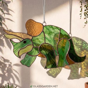 May include: A stained glass triceratops suncatcher featuring various shades of green and amber glass pieces held together by dark metal solder lines. The dinosaur-shaped ornament hangs from a thin white string and displays the text glassgarden.net.