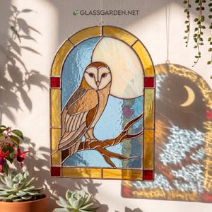 Puede incluir: Panel de vidrieras con un búho común posado en una rama sobre un fondo azul y crema. El panel tiene un borde dorado con detalles rojos y está suspendido por una cadena. Se ve la dirección del sitio web glassgarden.net.