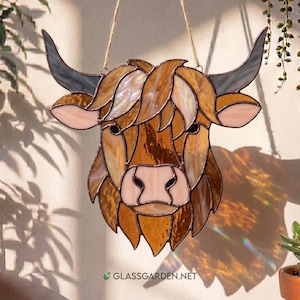 May include: A stained glass Highland cow head in shades of brown, tan, and orange. The artwork has a detailed design with textured glass, black eyes, and grey horns. A jute string is attached for hanging. The website address glassgarden.net is visible.
