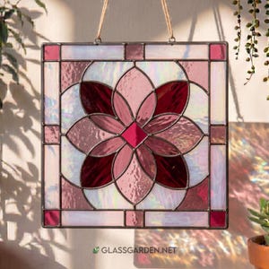 May include: A square stained glass panel featuring a floral design in pink and burgundy hues. The flower has a diamond-shaped centre and petal-shaped glass pieces. The panel is framed in dark metal and suspended by a natural fibre cord. The website address glassgarden.net is visible.