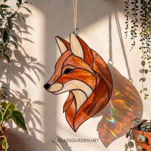 Puede incluir: Cabeza de zorro de vidriera en tonos naranja, marrón y crema, colgada de un lazo de cuerda. Esta pieza decorativa presenta un diseño detallado, ideal para añadir un toque de arte inspirado en la naturaleza a cualquier espacio. La dirección del sitio web es visible en la parte inferior.