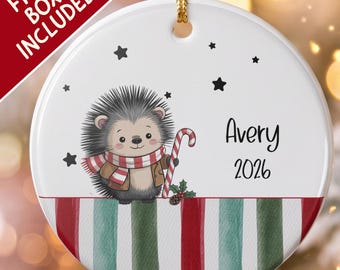 Pode incluir: Um ornamento redondo de cerâmica branca com um ouriço de desenho animado usando um cachecol de listras vermelhas e brancas e uma jaqueta marrom, segurando um bastão de doce. O texto Avery e 2026 está à direita. Listras vermelhas e verdes.
