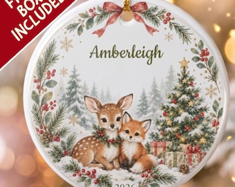 Pode incluir: Um enfeite de Natal redondo de cerâmica branca com uma ilustração impressa de um filhote de cervo e uma raposa sentados juntos na neve. O design inclui uma árvore de Natal decorada, ramos de pinheiro, bagas vermelhas e o nome Amberleigh com 2026.