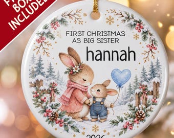 Pode incluir: Um ornamento redondo de cerâmica branca com uma cena de inverno de dois coelhos, um vestindo um casaco rosa e o outro jardineiras azuis, segurando um balão de coração azul. O texto diz FIRST CHRISTMAS AS BIG SISTER, hannah, 2026.