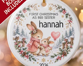 Pode incluir: Um ornamento redondo de cerâmica branca com uma cena de inverno de dois coelhos, um grande e um pequeno, com cachecóis cor-de-rosa. O texto diz FIRST CHRISTMAS AS BIG SISTER, hannah e 2026. Um balão rosa em forma de coração é segurado pelo coelho menor.