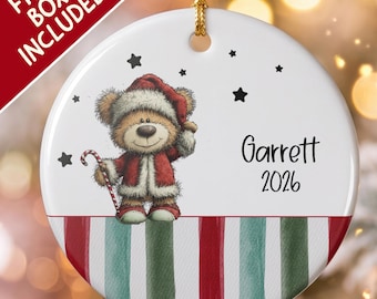 Pode incluir: Um enfeite de Natal redondo em cerâmica branca com um urso de peluche castanho a usar um gorro e casaco de Pai Natal vermelho, segurando um bastão de rebuçado. O enfeite apresenta o nome Garrett e o ano 2026, com riscas verticais.