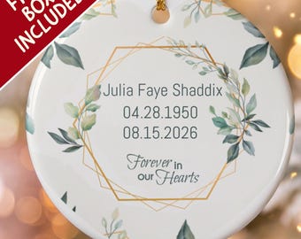 Pode incluir: Um ornamento redondo de cerâmica branca com uma moldura hexagonal geométrica dourada, folhas em aguarela verdes e o texto Julia Faye Shaddix, 04.28.1950, 08.15.2026 e Forever in our Hearts, suspenso por um cordão dourado.