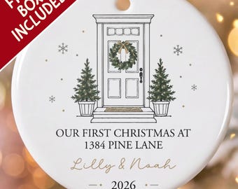 Pode incluir: Um ornamento redondo de cerâmica branca com um desenho de linhas pretas de uma porta da frente com uma guirlanda, ladeada por dois pinheiros em vasos. O texto diz: OUR FIRST CHRISTMAS AT 1384 PINE LANE, Lilly & Noah, e 2026. Inclui cordão.