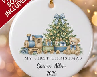 Pode incluir: Um ornamento redondo de cerâmica branca com um comboio azul, uma árvore de Natal e um urso de peluche. O texto diz MY FIRST CHRISTMAS, Spencer Allen e 2026. Um fio dourado está preso no topo para pendurar na árvore.