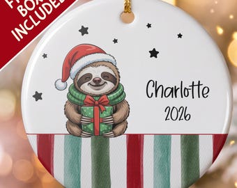 Pode incluir: Um ornamento redondo de cerâmica branca com um bicho-preguiça de desenho animado usando um gorro de Pai Natal vermelho e um cachecol verde, segurando um presente. Inclui estrelas pretas, o nome Charlotte e o ano 2026 sobre riscas.