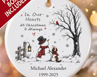 Pode incluir: Um ornamento redondo de cerâmica branca com um boneco de neve de cartola e cachecol xadrez vermelho, um cão, uma árvore nua com um cardeal vermelho, uma lanterna e flocos de neve. O texto diz In Our Hearts At Christmas & Always, Michael Alexander, 1999-2025.