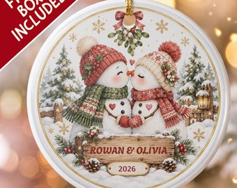 Pode incluir: Um enfeite de Natal redondo em cerâmica branca com dois bonecos de neve usando gorros e cachecóis de tricô, beijando-se sob o visco. Uma placa de madeira exibe o texto ROWAN & OLIVIA e o ano 2026 em um cenário de floresta nevada.