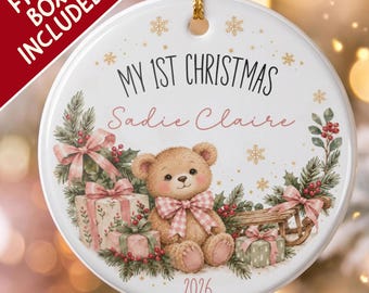 Pode incluir: Um enfeite de Natal redondo de cerâmica branca com um urso de peluche castanho com um laço xadrez rosa, rodeado de presentes, ramos de pinheiro e bagas vermelhas. O texto diz MY 1ST CHRISTMAS, Sadie Claire e 2026.
