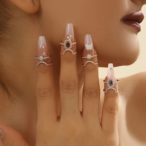 May include: Four silver-toned rings featuring clear cubic zirconia stones and central rectangular or circular settings. Each ring is worn on a finger with long, light pink coffin-shaped nails decorated with white star and butterfly accents.