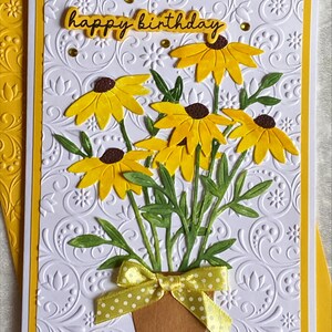 May include: A handmade birthday card featuring a bouquet of yellow flowers with brown centers in a brown pot, tied with a yellow polka dot bow. The card has a white embossed background and the words "happy birthday" in yellow.